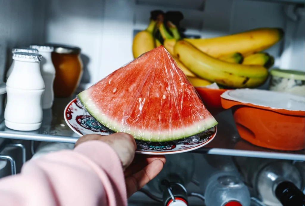 Como armazenar frutas cortadas na geladeira e evitar que estraguem rápido 2 Uma mão segura um prato com uma fatia de melancia embalada em plástico filme dentro de uma geladeira. Ao fundo, veem-se bananas e garrafas de iogurte.