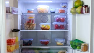 Home 1 Interior de uma geladeira aberta e organizada, com prateleiras cheias de potes transparentes com alimentos e uma gaveta com vegetais frescos.