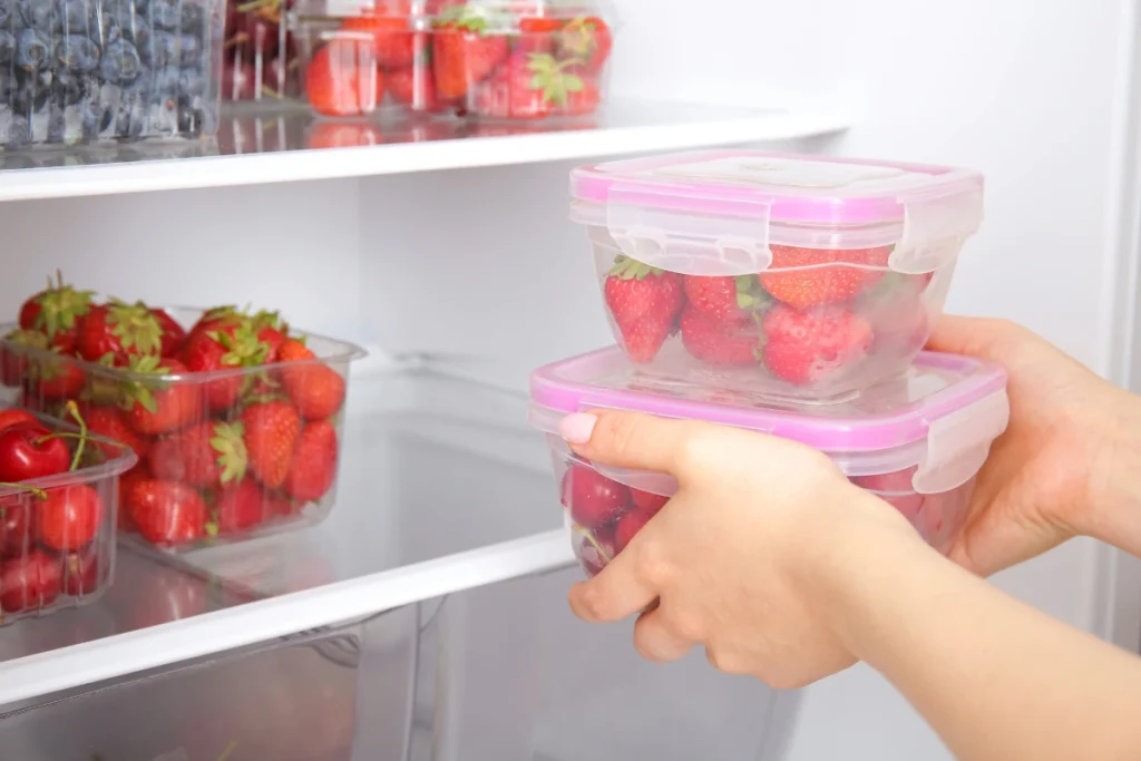 Como armazenar frutas cortadas na geladeira e evitar que estraguem rápido 1 Close de uma pessoa organizando frutas vermelhas frescas em potes herméticos dentro da geladeira, destacando o armazenamento correto e a conservação dos alimentos.