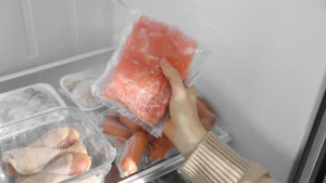 Close de uma pessoa organizando carnes e aves congeladas em recipientes e sacos plásticos dentro do freezer, destacando o armazenamento correto dos alimentos.