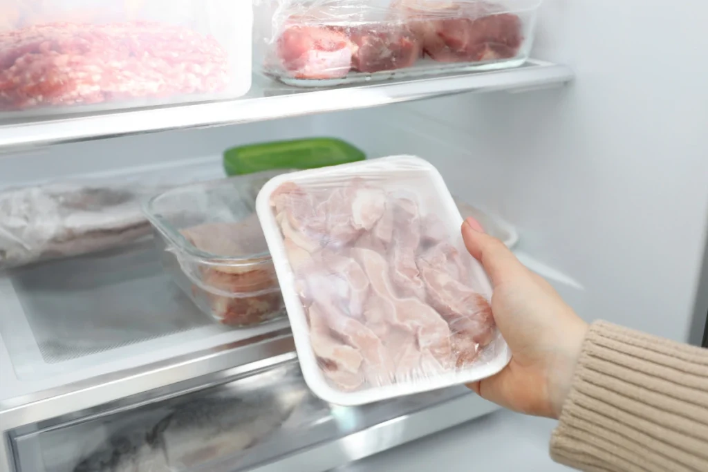 Mão de uma pessoa segurando uma bandeja de carne embalada com plástico filme, retirando-a de dentro de uma geladeira organizada.