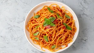 Home 1 Prato branco redondo com macarrão espaguete ao molho de tomate fresco, decorado com folhas de manjericão e metades de tomate-cereja, visto de cima.