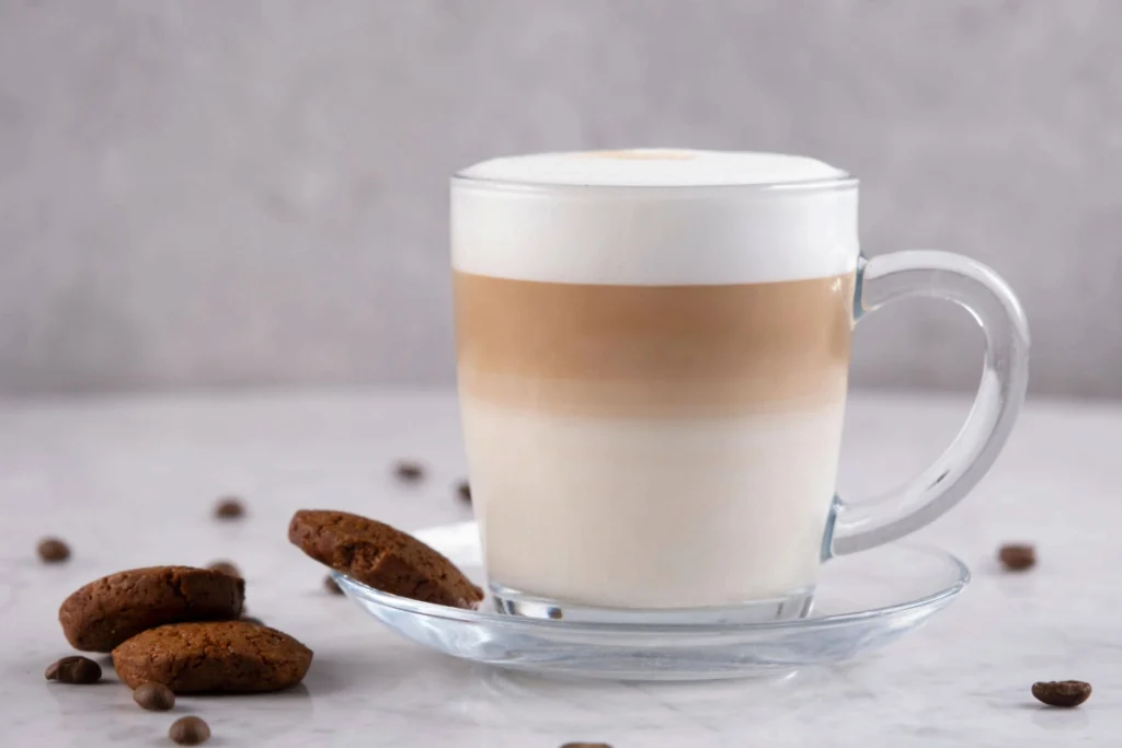 Xícara de vidro com café latte em camadas, acompanhada de biscoitos de chocolate e grãos de café sobre superfície de mármore clara.