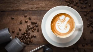 Xícara de café com latte art acompanhada de grãos de café torrados e moedor sobre mesa de madeira, ideal para um café especial Cocamar.