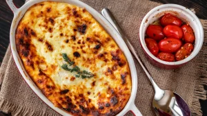 Foto aérea de um prato de arroz de forno cremoso e gratinado em um refratário oval, acompanhado por uma tigela com tomates cereja assados e uma colher de servir, sobre um jogo americano de juta em mesa de madeira.