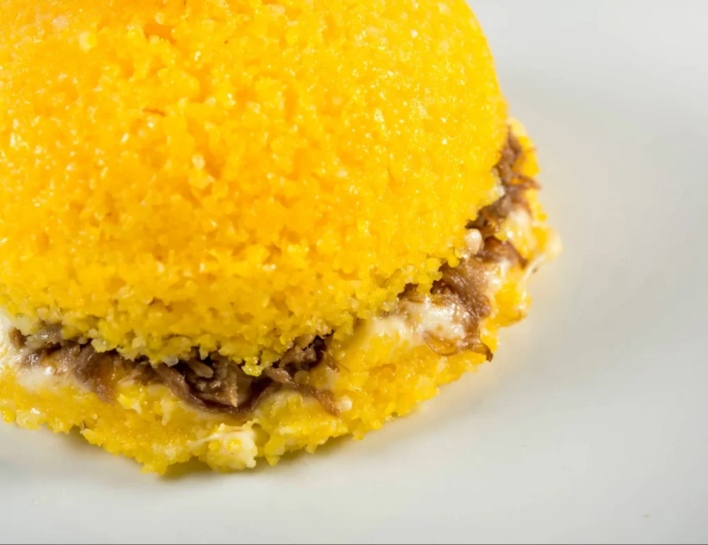 Close-up de uma porção de cuscuz amarelo recheado com carne seca desfiada e queijo cremoso derretido, sobre fundo branco. A textura do cuscuz é bem granulada e úmida.