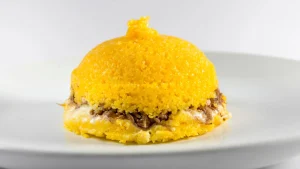 Home 1 Cuscuz amarelo servido em formato cônico sobre um prato branco, recheado no centro com carne-seca desfiada e queijo derretido.