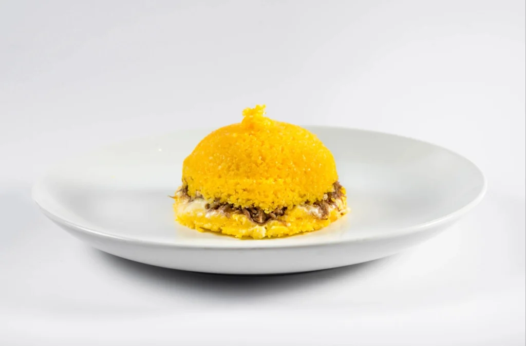Vista frontal de um prato raso branco centralizado em fundo infinito branco, contendo uma porção individual de cuscuz amarelo cônico, recheado com carne-seca desfiada e queijo derretido.