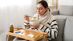 Mulher resfriada, enrolada em um cachecol e suéter, sentada no sofá comendo sopa quente com uma colher, enquanto uma bandeja de madeira na sua frente exibe remédios e lenços de papel.