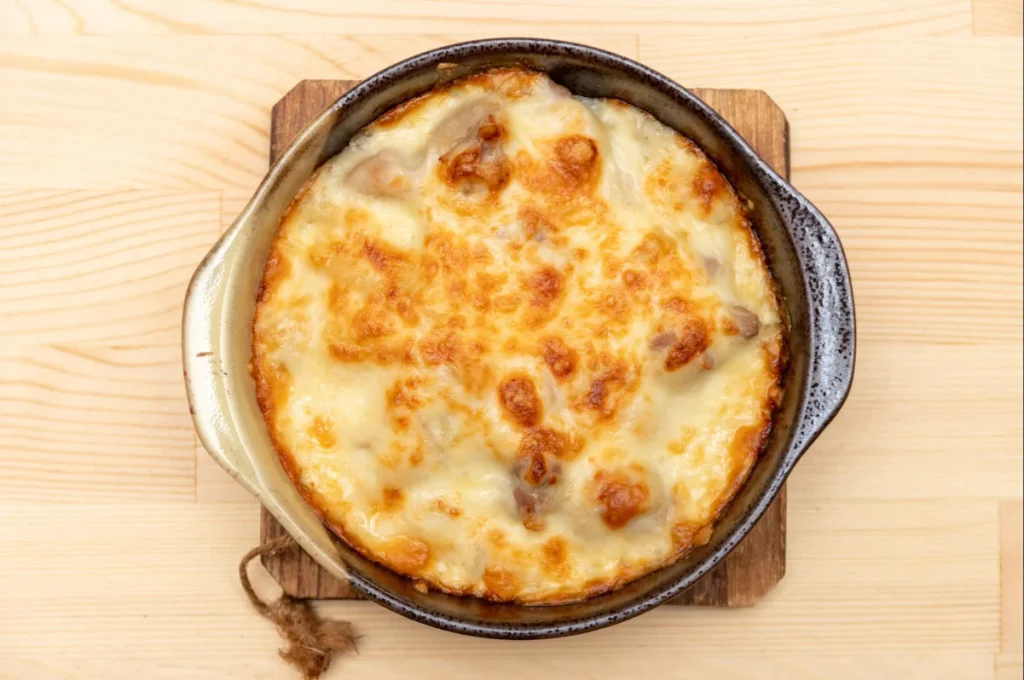 Vista superior de uma travessa redonda de cerâmica com arroz de forno cremoso e cobertura de queijo gratinado dourado, servida sobre um descanso de madeira em uma mesa clara.
