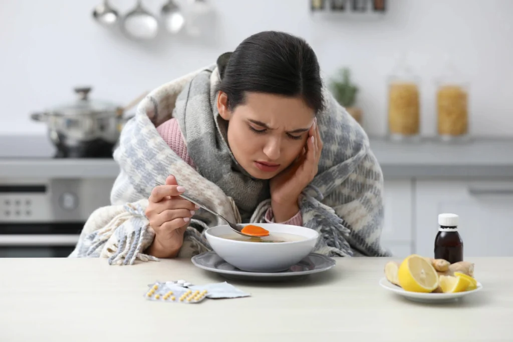  Mulher doente enrolada em um cobertor xadrez, sentada à mesa da cozinha tomando sopa quente com legumes, com a mão na cabeça demonstrando indisposição, ao lado de remédios em cartela, xarope e pedaços de limão e gengibre.