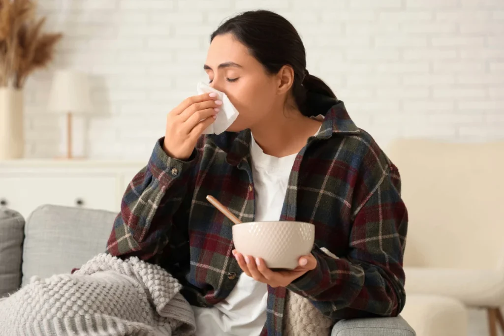 Mulher gripada usando uma camisa xadrez, segurando uma tigela de sopa quente em uma das mãos enquanto assoa o nariz com um lenço de papel, sentada no sofá de casa.